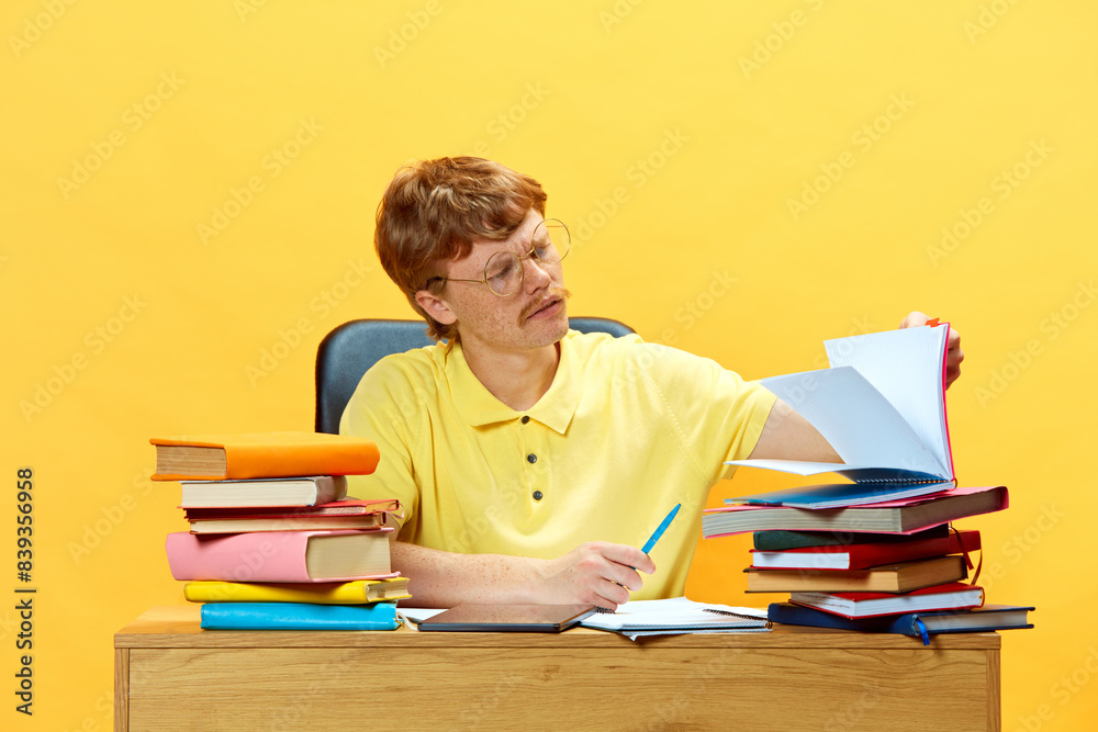 Young man, student sitting at desk and doing homework surrounded huge stack of books against vibrant yellow background. Concept of education and study, skills, knowledges, graduation. Ad Young man, student sitting at desk and doing homework surrounded huge stack of books against vibrant yellow background. Concept of education and study, skills, knowledges, graduation. Ad