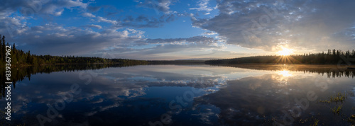 A peaceful morning panorama at a calm northern lake with the sun just rising above a distant treeline. The calm water makes a perfect mirror showing the clouds in a blue sky. 