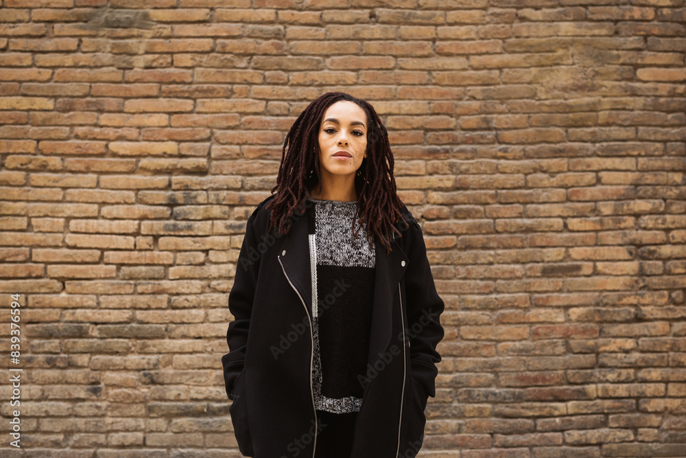 Portrait of a young beautiful woman with dreadlocks standing against a brick wall on the street.