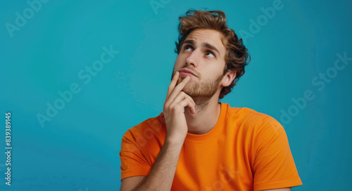 A woman in an orange shirt is looking confused and thinking with his hand on chin against blue background © Kien