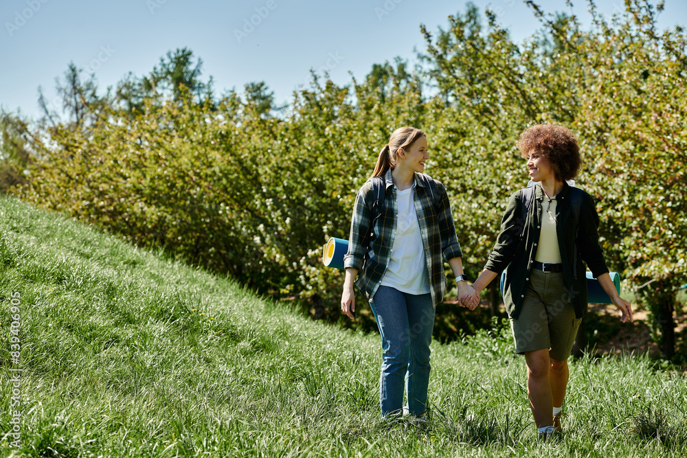 Obraz premium A young, multicultural lesbian couple hikes hand-in-hand through a lush green meadow, enjoying a day outdoors.