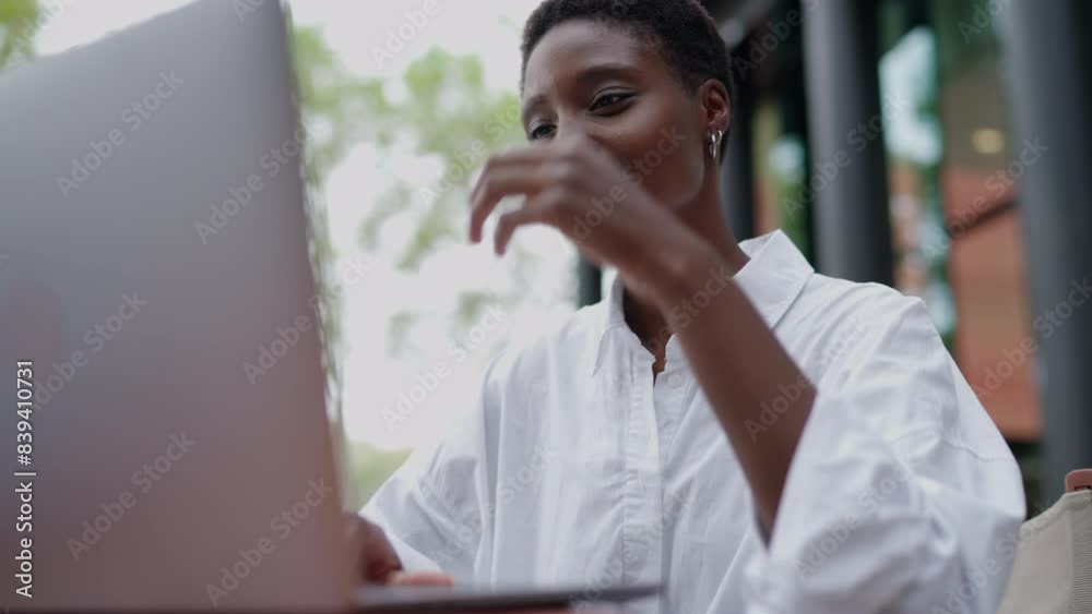 Cheerful young Black woman working as digital nomad, types on laptop computer at outdoor café. Engaged and smiling female student is dressed in white shirt, enjoy writing article studying project
