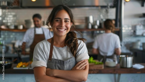 A smiling female restaurant owner standing in her kitchen with arms crossed. People working behind the counter out of focus in the background
