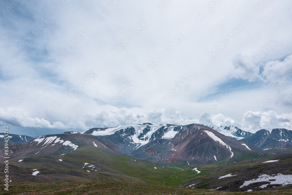Dramatic view to green alpine valley with long creek and large snow-capped mountain range under cloudy gray sky. Mountain stream among rock hills with snowfields and high mountains. Awesome landscape. Dramatic view to green alpine valley with long creek and large snow-capped mountain range under cloudy gray sky. Mountain stream among rock hills with snowfields and high mountains. Awesome landscape.