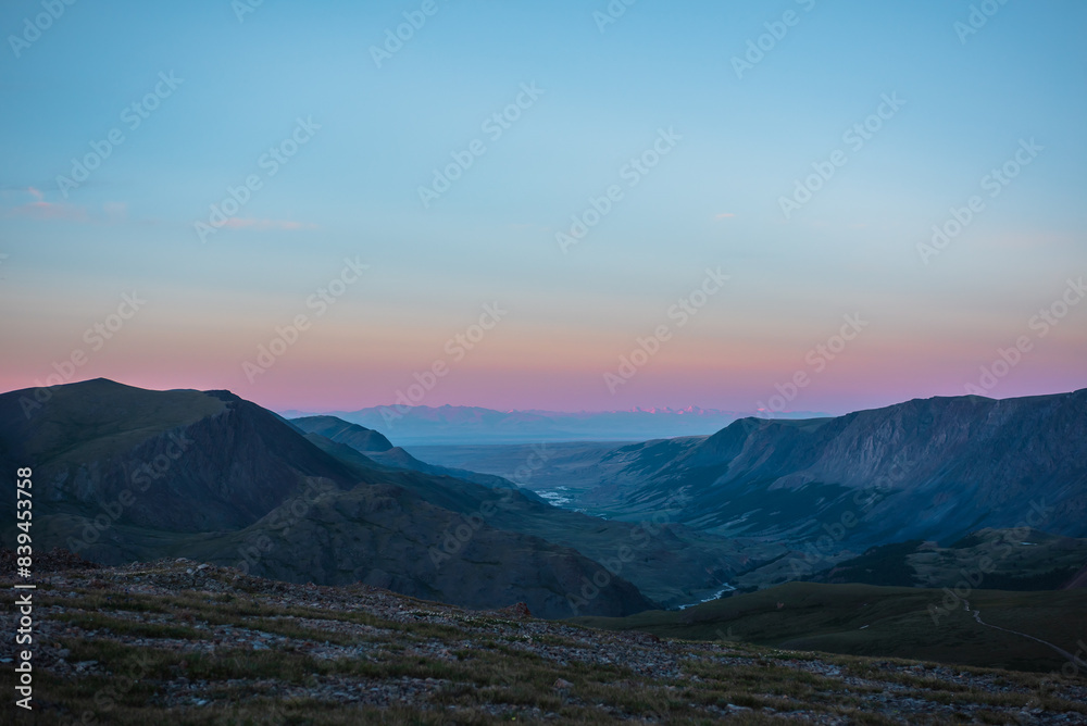 Top view from pass to wide alpine valley with long river in shadow against large mountain range silhouette with snow-capped peaks of vivid pink color far away at sunset. Big snowy ridge on horizon.