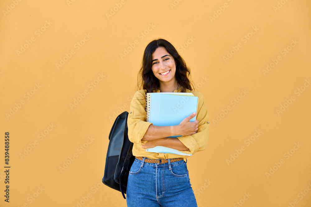© Stock 4 You - Young Arabian or Indian holding books, notebooks, ready for studying. Smiling portrait of beautiful middle eastern Israel girl student with backpack isolated on yellow background looking at camera.