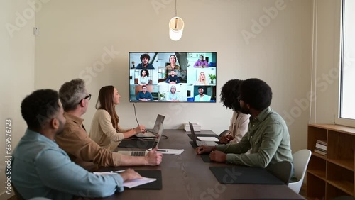 Team engaged in a virtual meeting with a display screen, showing both remote participants and in-person colleagues interacting seamlessly, integration of modern technology in collaborative workspaces