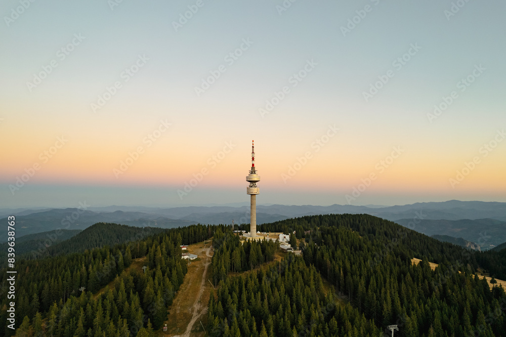 Obraz premium Aerial view tower structure high in mountains aerial view drone forward, dense pine forest at sunrise in summer morning on blue sky. Panorama post-dawn blue sky high in mountains. Nature Bulgaria
