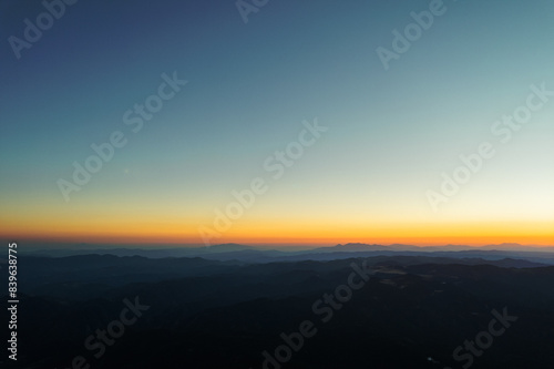 Sunset cloudy sky evening horizon from aerial view. Lens flare in mountains at orange sunset in summer morning aerial view drone. Sun rays. Green pine forest slopes of mountain range. Bright disk sun