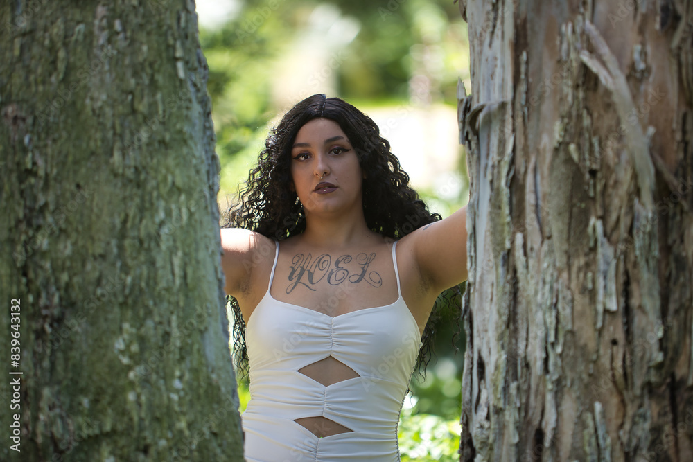 Young, beautiful, alternative, chubby woman with tattoos, wearing a white dress, sad and alone, looking at the camera, leaning between the trunks of a big tree. Millennial concept, plump, different. Young, beautiful, alternative, chubby woman with tattoos, wearing a white dress, sad and alone, looking at the camera, leaning between the trunks of a big tree. Millennial concept, plump, different.
