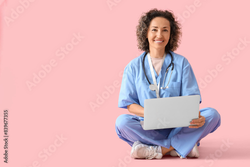 Mature female doctor working with laptop on pink background