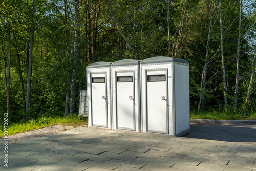 A row of three white portable toilets stand in a clearing in a wooded area photo