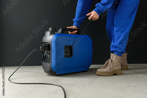 A woman in a blue suit starts a generator. A modern portable gasoline inverter generator stands in a garage against a black wall. Backup autonomous power supply at home. Electricity production.
