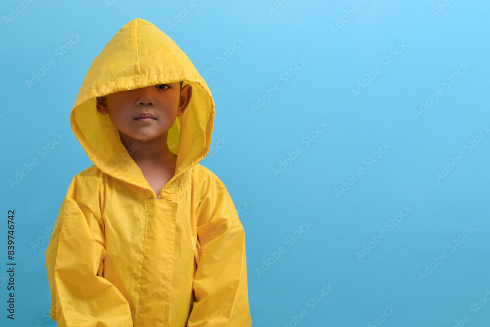 Cute little Asian boy wearing a yellow raincoat isolated on blue background. Weather forecast concept Cute little Asian boy wearing a yellow raincoat isolated on blue background. Weather forecast concept