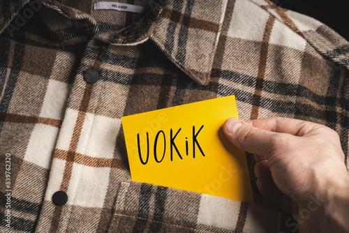 Yellow card with a handwritten inscription "UOKiK", held in the hand against the background of a brown plaid shirt (selective focus)