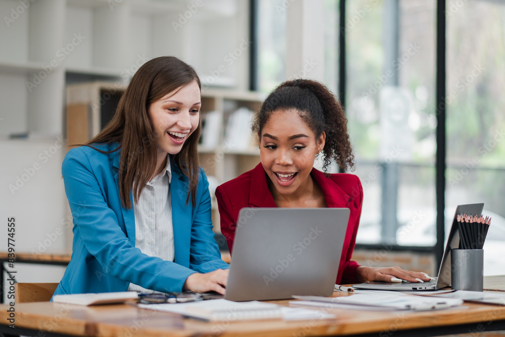Two women are sitting at a desk with a laptop open in front of them. They are smiling and seem to be having a good time Two women are sitting at a desk with a laptop open in front of them. They are smiling and seem to be having a good time