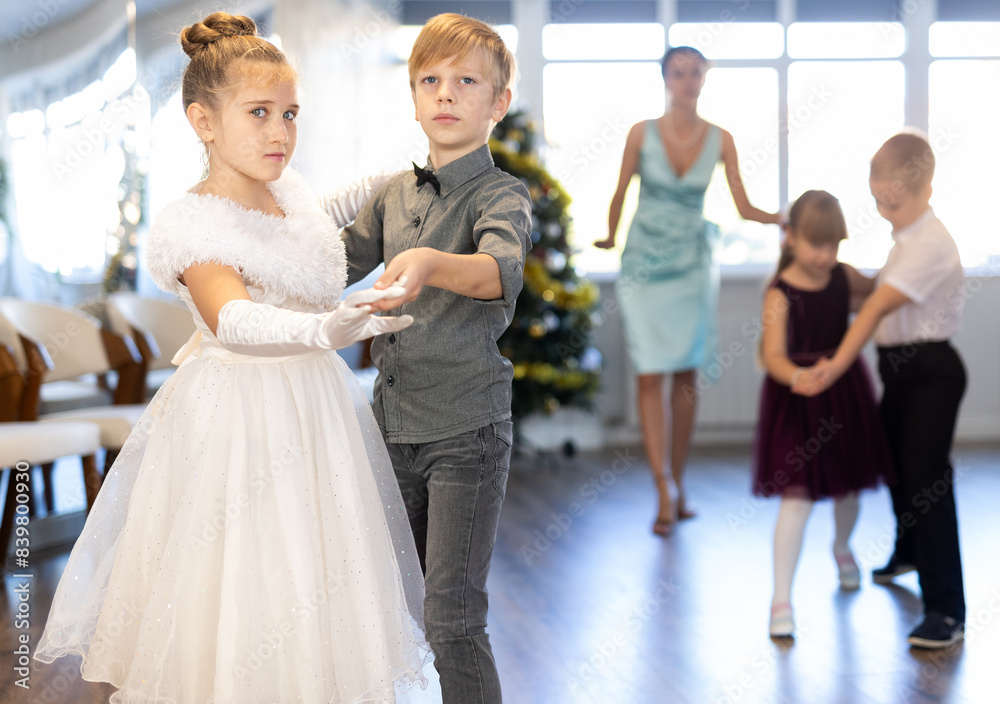 Obraz premium Positive elementary students in festive attire rehearsing ballroom dance number for upcoming Christmas party at school with interested female teacher in background..