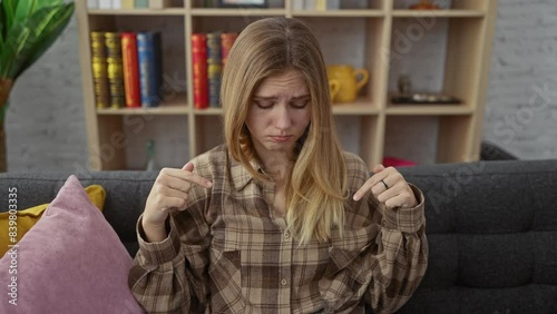Young blonde woman, upset and unhappy, sitting sullenly on sofa indoors, pointing down with a sad look, indicating direction depressively. depressed at home, a poignant portrait of desmotivation.