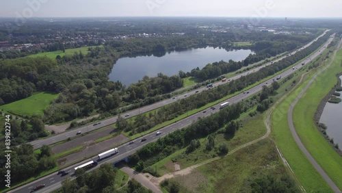 Stunning aerial footage capturing the E19 highway between Brussels and Antwerp near Mechelen. The scene features a tranquil lake adjacent to the highway, with lush greenery and forested areas photo