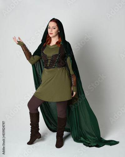 Full length portrait of red haired female model wearing green medieval fantasy costume leather armour, dramatic long flowing hooded velvet cape. Standing walking pose, isolated white studio background