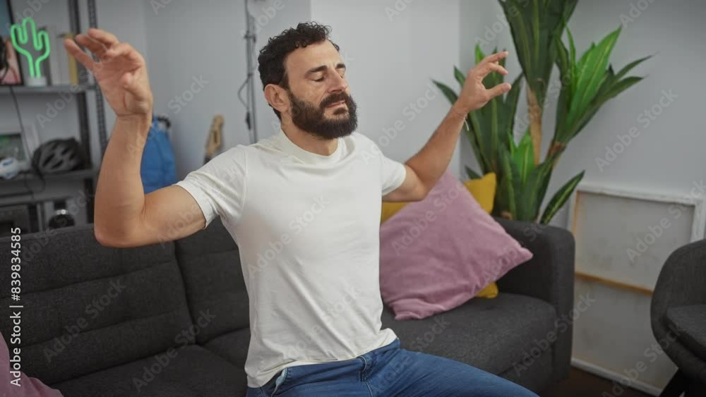 Attractive middle-aged man, hands posed in peaceful yoga meditation, radiates calmness as he closes his eyes and relaxes in the comfort of his home, wearing a simple white t-shirt.
