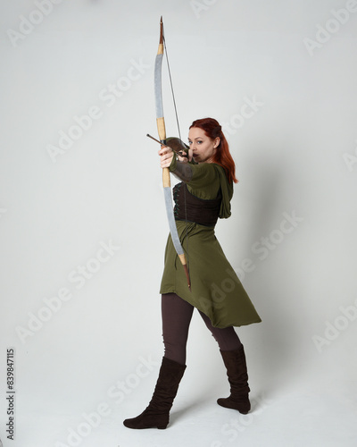 Full length portrait of red haired female model wearing green medieval fantasy costume, leather armour.
Holding archery bow and arrow weapon, standing action pose isolated on white studio background.
