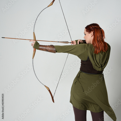 close up portrait of red haired female model wearing green medieval fantasy costume, leather armour. Holding archery bow and arrow weapon, standing action pose isolated on white studio background.