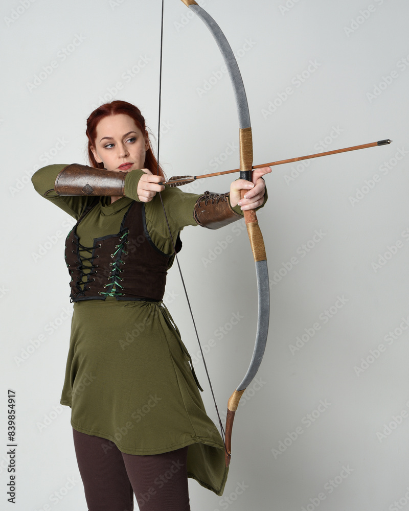 close up portrait of red haired female model wearing green medieval fantasy costume, leather armour.
Holding archery bow and arrow weapon, standing action pose isolated on white studio background. close up portrait of red haired female model wearing green medieval fantasy costume, leather armour.
Holding archery bow and arrow weapon, standing action pose isolated on white studio background.