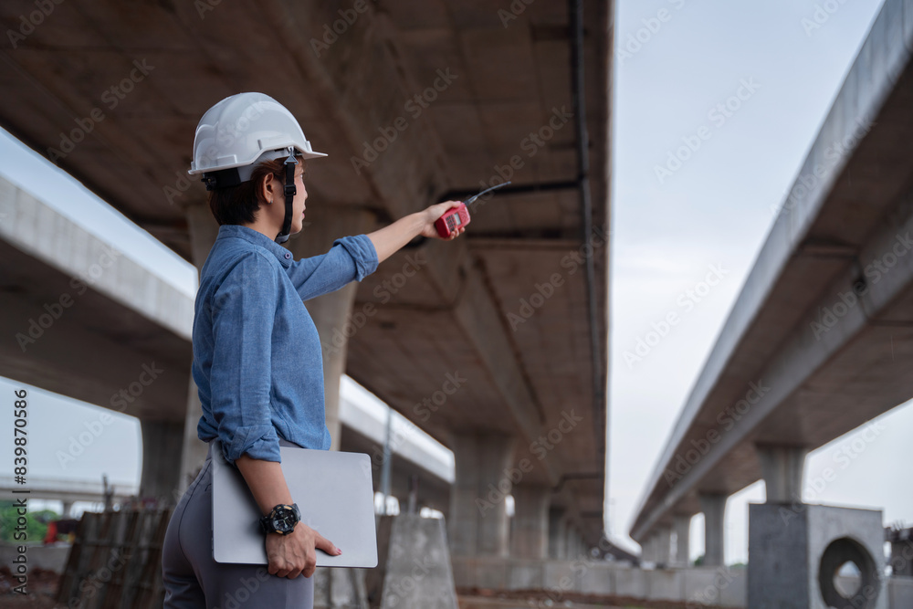 Asian female engineer supervisor under the overpass construction, using a laptop to analyze data and coordinate with her team, demonstrating leadership.