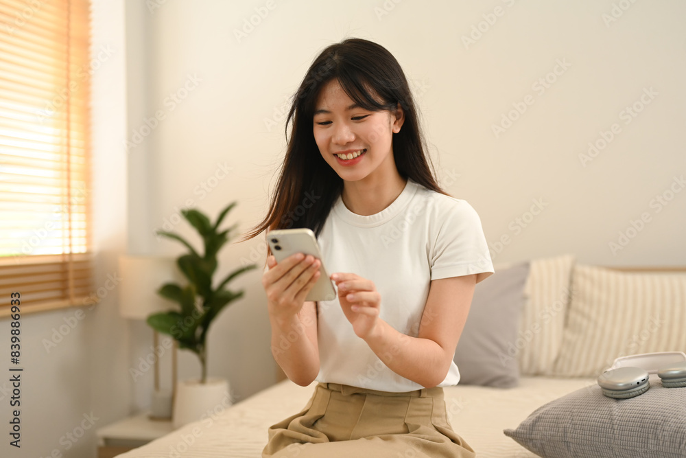 Pretty young woman using mobile phone while sitting on bed at home Pretty young woman using mobile phone while sitting on bed at home