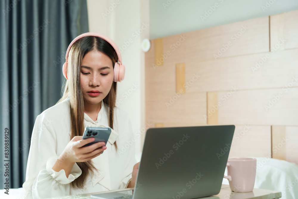 Asian woman using smartphone while working on laptop with pink headphones. Casual attire, long hair. Modern home office setup, beige decor, natural light. Focused expression, pink coffee cup on table. Asian woman using smartphone while working on laptop with pink headphones. Casual attire, long hair. Modern home office setup, beige decor, natural light. Focused expression, pink coffee cup on table.