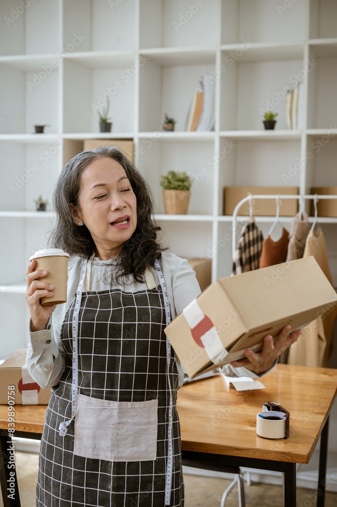 A happy mature Asian female online seller is holding a coffee cup and a cardboard box in her office. A happy mature Asian female online seller is holding a coffee cup and a cardboard box in her office.
