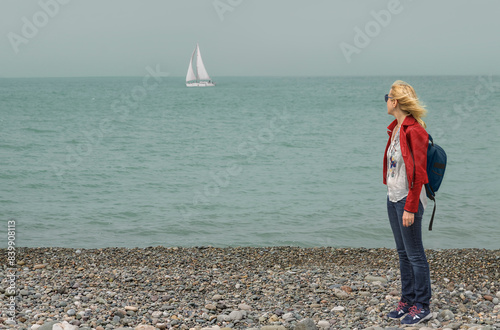 Принти на полотні A woman in a red jacket and with a backpack stands on a pebble beach and looks at a sailboat in the sea