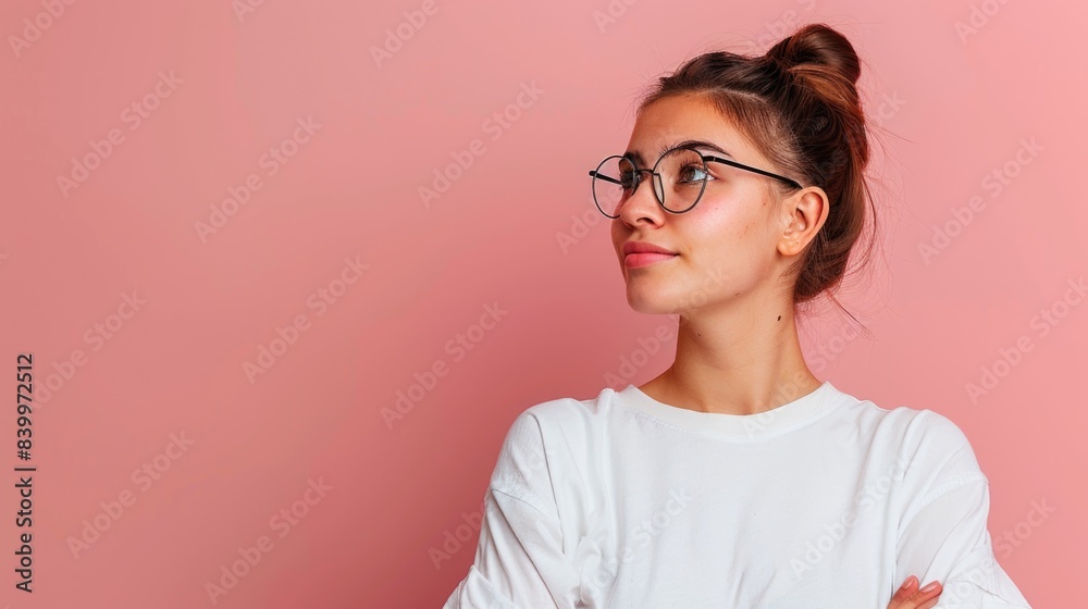 A young businesswoman wearing glasses looks to the side with a thoughtful expression while standing in front of a pink background A young businesswoman wearing glasses looks to the side with a thoughtful expression while standing in front of a pink background