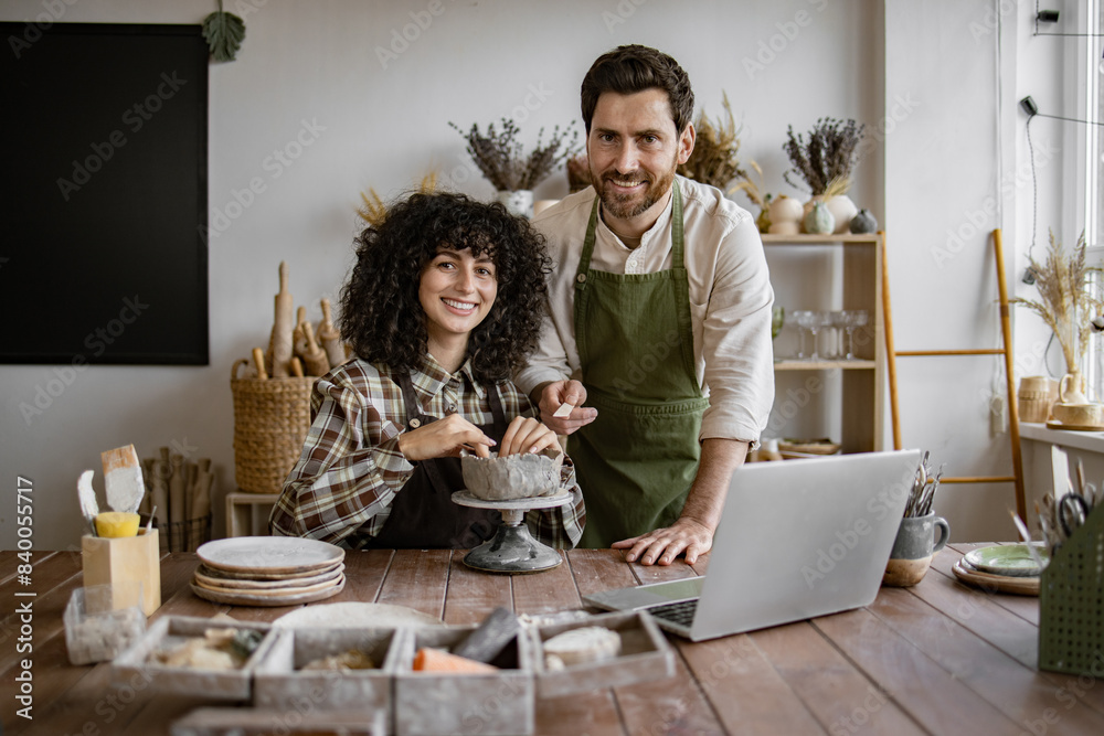 Obraz premium Smiling couple working together on pottery project in art studio. Creative process with laptop and ceramic tools on wooden table.