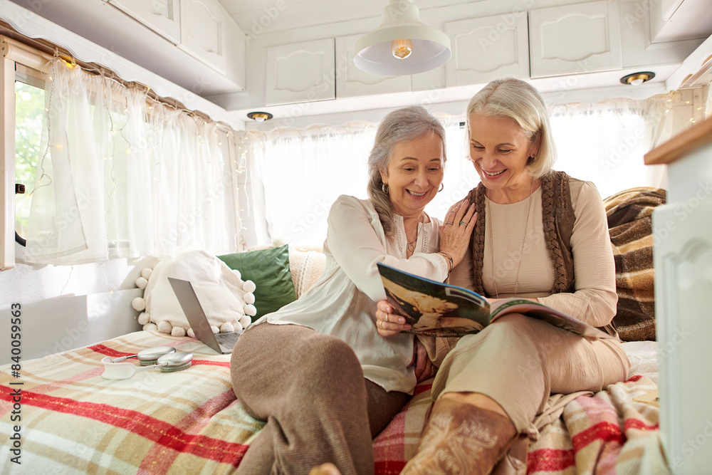 Two middle-aged women, a lesbian couple, are happily relaxing in their camper van during a trip. They are sitting on the bed, reading a magazine. Two middle-aged women, a lesbian couple, are happily relaxing in their camper van during a trip. They are sitting on the bed, reading a magazine.