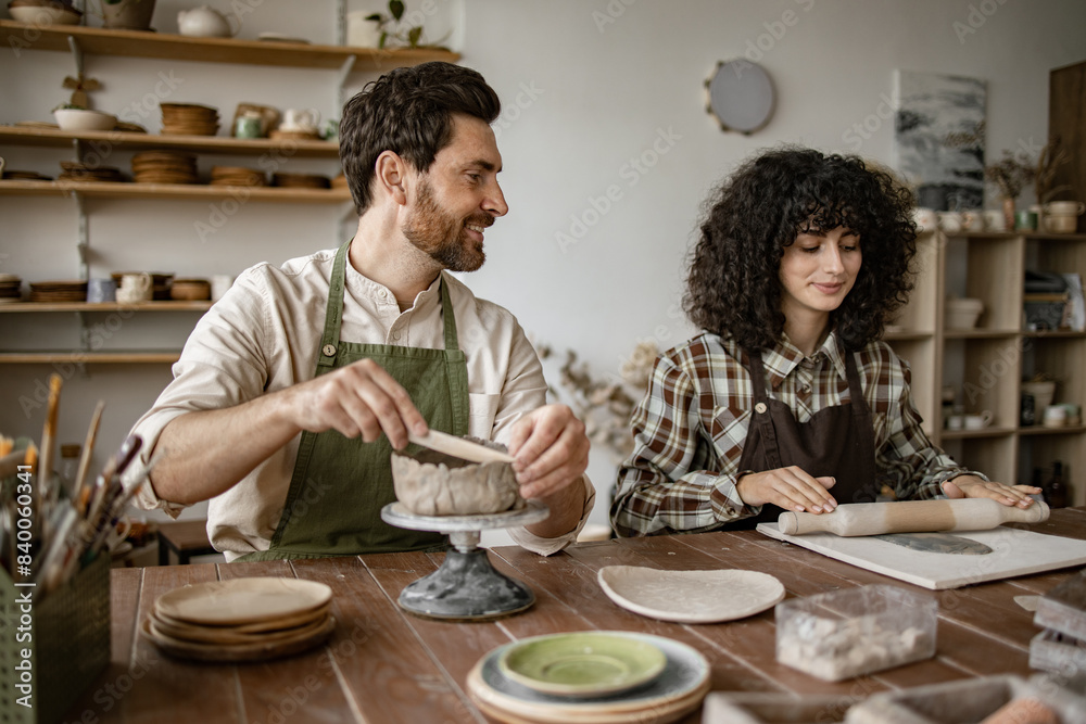 Bearded mature man in apron sculpts vase while woman rolls clay with rolling pin during pottery class in studio. Bearded mature man in apron sculpts vase while woman rolls clay with rolling pin during pottery class in studio.