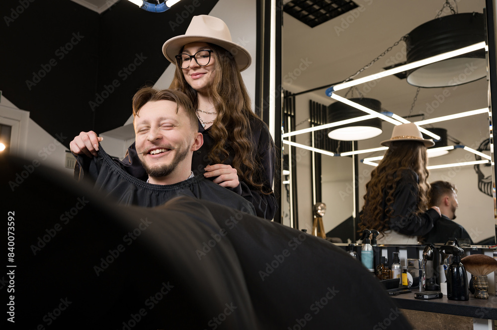 Obraz premium A young man with a beard in a black cape sits on a chair in a barbershop. The client is waiting for a haircut of the beard.