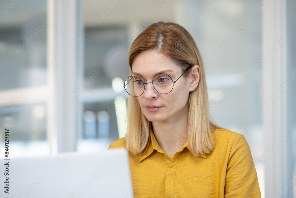 Focused woman wearing glasses working on a laptop in a modern office environment. Concentration, productivity, and dedication in a professional workspace.
