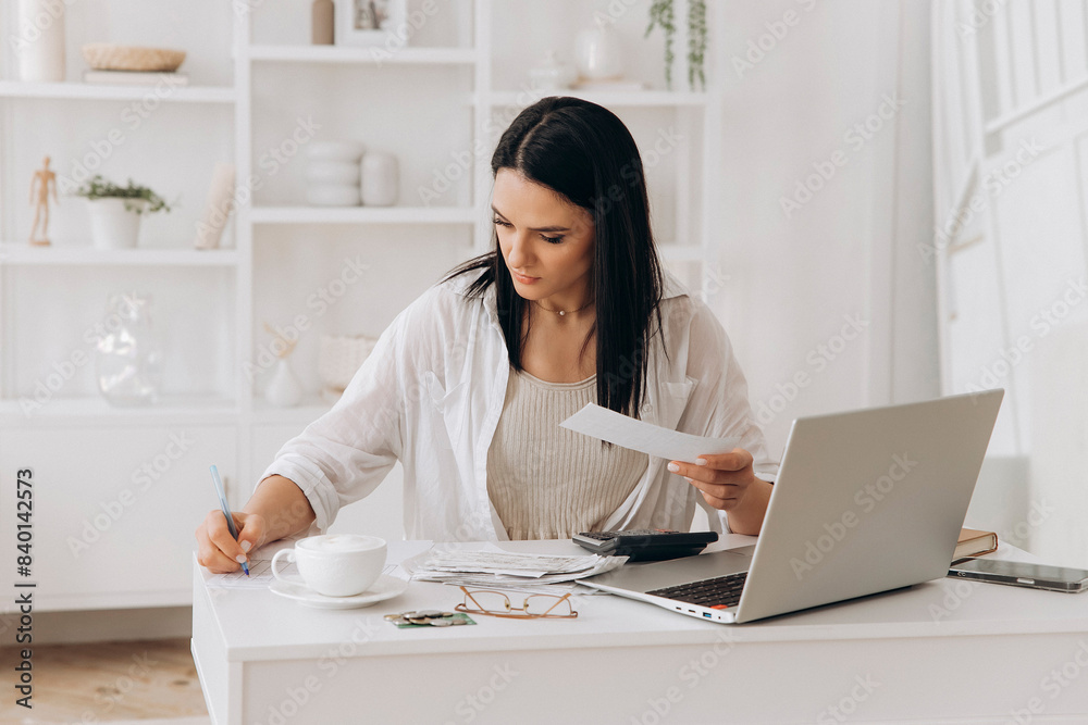 Confident young woman working at desk with laptop, documents, and calculator. Writing notes while reviewing papers. Managing utility payments, taxes, invoices in home office with white shelves Confident young woman working at desk with laptop, documents, and calculator. Writing notes while reviewing papers. Managing utility payments, taxes, invoices in home office with white shelves