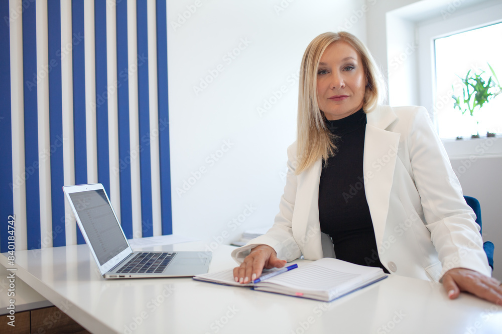 A beautiful middle aged lady is dressed in a strict suit, a white jacket  and a black dress/ The woman sits at the workplace with a computer and works