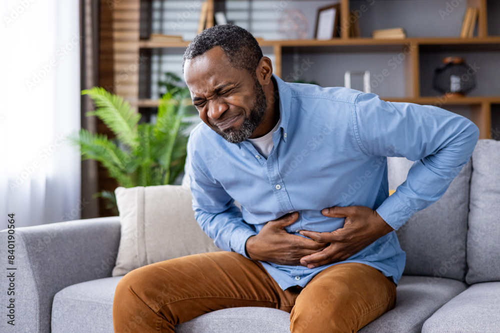 Man in discomfort, holding his stomach while sitting on a sofa, indicating possible pain or indigestion in a home setting. Man in discomfort, holding his stomach while sitting on a sofa, indicating possible pain or indigestion in a home setting.