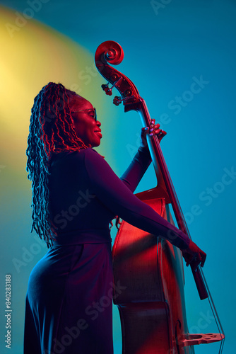 Canvas Print Young African-American woman, musician performing, playing on double bass in neon light against colorful gradient background