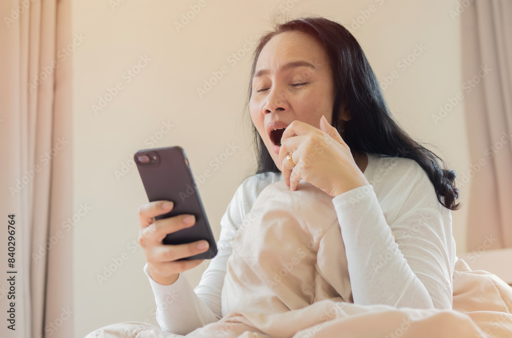 Shot image of mature woman using mobile phone in bed at morning while yawning and still sleepy. Shot image of mature woman using mobile phone in bed at morning while yawning and still sleepy.