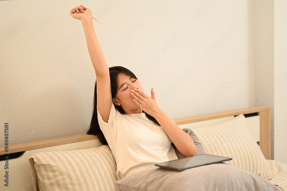 Sleepy young woman yawning and covering mouth with hand while sitting with digital tablet in bed Sleepy young woman yawning and covering mouth with hand while sitting with digital tablet in bed