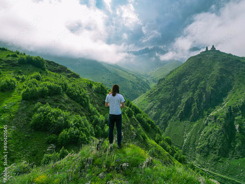long-haired man stands on the top of a green mountain in a white shirt - drone shot