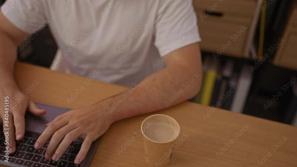 A young man working on a laptop in an organized office or indoor workplace, using technology with a beverage cup on the desk. A young man working on a laptop in an organized office or indoor workplace, using technology with a beverage cup on the desk.