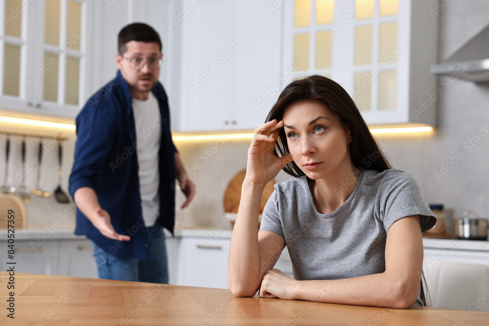 Stressed wife sitting at table while her angry husband screaming at her in kitchen, selective focus. Relationship problems Stressed wife sitting at table while her angry husband screaming at her in kitchen, selective focus. Relationship problems