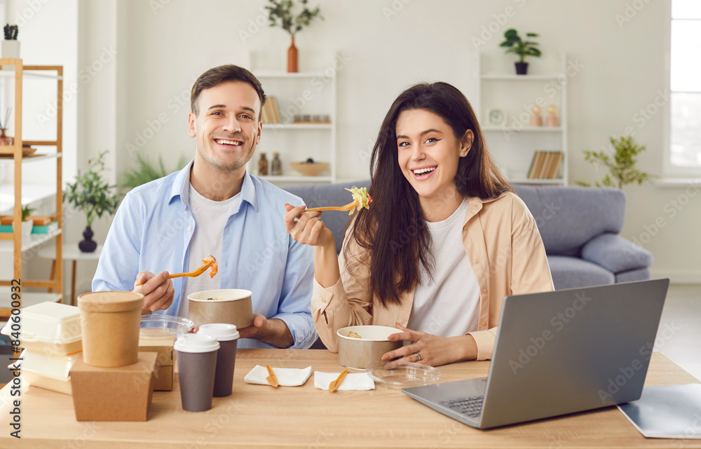 Portrait of happy young married couple sitting at table with laptop and eating fresh fast food in boxes having a lunch together making online order. Take away food and delivery service concept. Portrait of happy young married couple sitting at table with laptop and eating fresh fast food in boxes having a lunch together making online order. Take away food and delivery service concept.