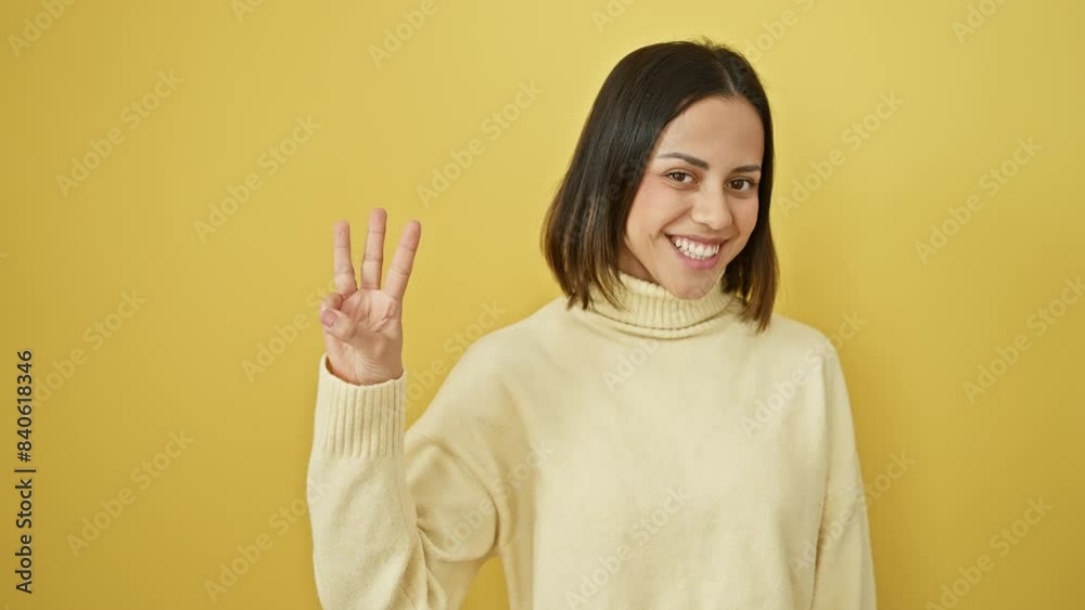 Cheerful young hispanic woman in yellow backdrop, smiling confidently, raising three fingers in a gesture of showing 'number three'. wearing a stylish sweater, a picture of beauty.